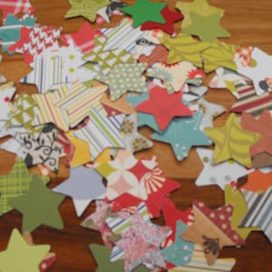 Puede incluir: Una colección de estrellas de papel de colores con varios patrones y diseños. Las estrellas están hechas de diferentes materiales, incluyendo papel estampado, tela y cartulina. Las estrellas están esparcidas sobre una superficie de madera.