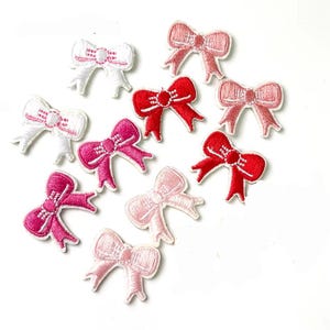 May include: Assortment of ten embroidered iron-on patches in the shape of bows. The bows are in shades of pink, red, and white.