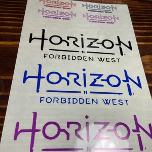 Puede incluir: Cuatro pegatinas con el texto "Horizon Forbidden West" en diferentes colores y fuentes. Las pegatinas est&aacute;n sobre una superficie de madera marr&oacute;n claro.