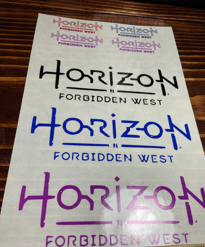 Puede incluir: Cuatro pegatinas con el texto "Horizon Forbidden West" en diferentes colores y fuentes. Las pegatinas est&aacute;n sobre una superficie de madera marr&oacute;n claro.