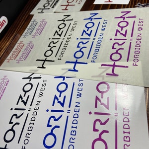 Puede incluir: Cuatro pegatinas de vinilo con el texto "Horizon Forbidden West" en diferentes colores y fuentes. Las pegatinas est&aacute;n sobre una superficie de madera.