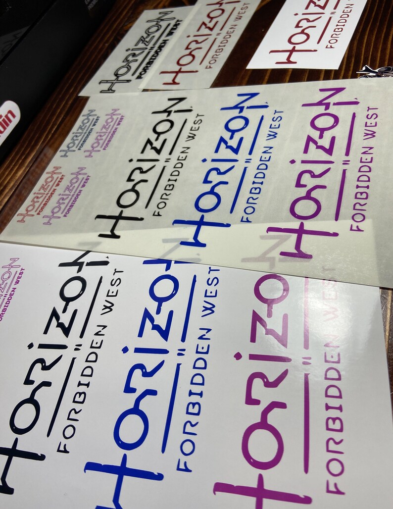 Puede incluir: Cuatro pegatinas de vinilo con el texto "Horizon Forbidden West" en diferentes colores y fuentes. Las pegatinas est&aacute;n sobre una superficie de madera.