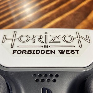 Puede incluir: Un mando de PlayStation blanco con una pegatina negra que dice "Horizon Forbidden West".