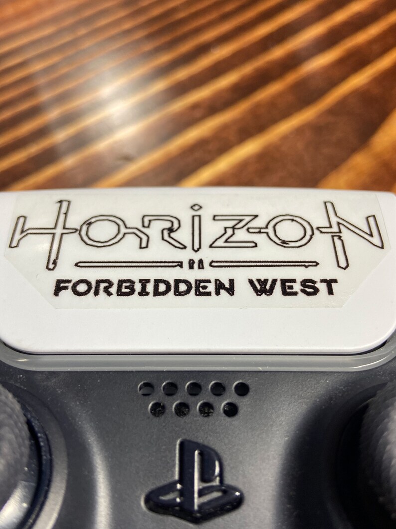 Puede incluir: Un mando de PlayStation blanco con una pegatina negra que dice "Horizon Forbidden West".