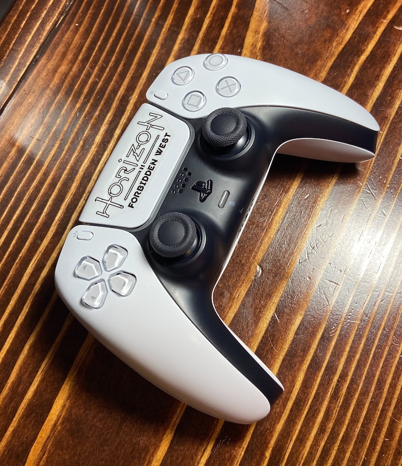 Puede incluir: Un mando de juego PlayStation 5 DualSense blanco y negro con una piel personalizada que presenta el texto "Horizon Forbidden West".