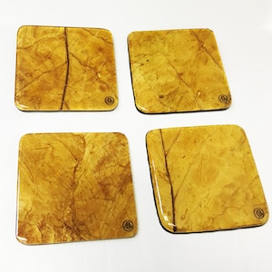May include: Set of four square coasters with a marbled, yellow and brown design. Each coaster has a small, circular logo in the bottom right corner.