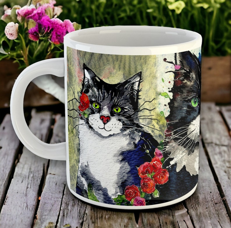 A large white ceramic mug decorated with 3 watercolour paintings of black and white cats, surrounded by red and pink flowers.