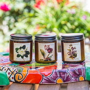 May include: Three amber glass candle jars with lids, each featuring a different floral illustration and the text "Flower Garden". The candles are arranged on a colorful, circular, ceramic tile.