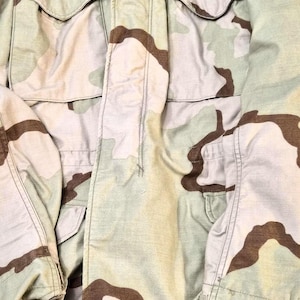 May include: A tan and brown camouflage military jacket with multiple pockets and a zipper closure. The jacket has a button-down front and a collar. The fabric appears to be a durable, heavy-duty material.