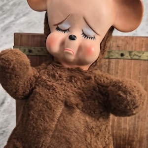 Può includere: Un giocattolo di peluche vintage raffigurante un orso con un'espressione triste, grandi orecchie e un corpo di pelliccia marrone. L'orso ha un viso dipinto con occhi chiusi e guance rosee. È posizionato davanti a una struttura in legno.