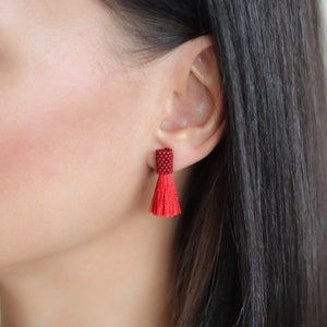 Peut inclure: Une paire de boucles d'oreilles à pompon rouge avec un dessus en perles rouge foncé. Les boucles d'oreilles sont portées par une personne aux cheveux foncés.
