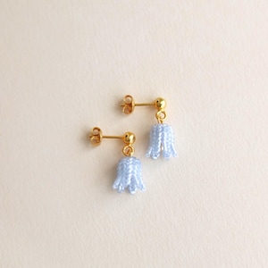 Può includere: Un paio di orecchini a bottone color oro con pendenti a forma di campana azzurra. Gli orecchini sono su uno sfondo color crema. Un accessorio delicato di ispirazione floreale.