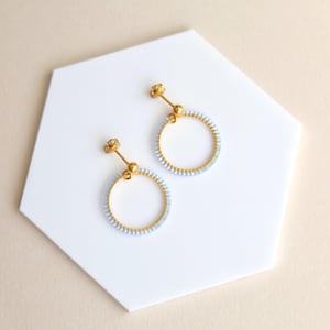 Può includere: Un paio di orecchini a cerchio color oro con un design di perline azzurre. Gli orecchini sono esposti su una superficie esagonale bianca su uno sfondo neutro. Gli orecchini hanno una chiusura a perno.