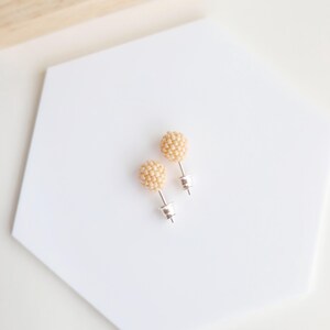 May include: A pair of stud earrings featuring a cluster of small, light beige beads. The earrings have silver-colored posts and backs. The earrings are displayed on a white, hexagonal surface.