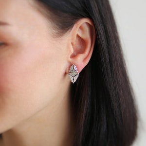 May include: A close-up of a woman's ear with a pair of silver geometric earrings. The earrings are shaped like a diamond and have a textured surface.