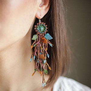 May include: A pair of long, beaded earrings with a floral design. The earrings are made with brown, green, and blue beads and feature a small, round, iridescent stone in the center of each flower.