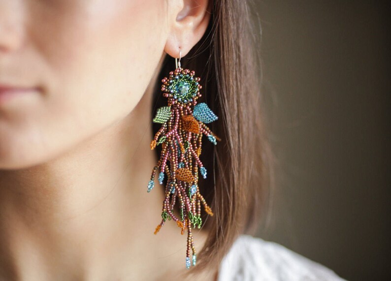 May include: A pair of long, beaded earrings with a floral design. The earrings are made with brown, green, and blue beads and feature a small, round, iridescent stone in the center of each flower.
