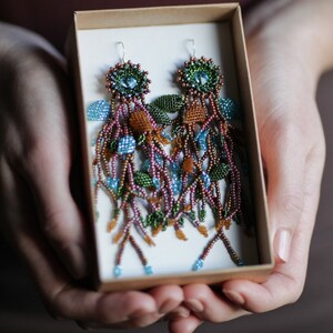 May include: A pair of long, beaded earrings in a box. The earrings are made with brown, green, and blue beads and have a floral design.