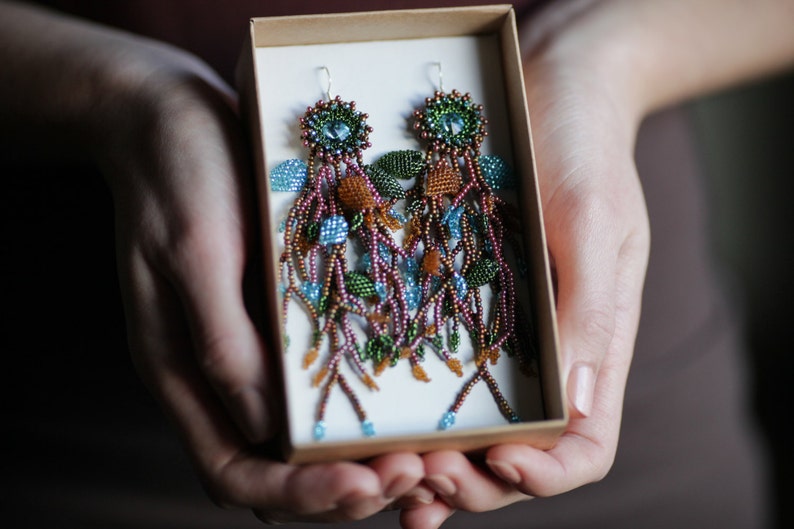 May include: A pair of long, beaded earrings in a box. The earrings are made with brown, green, and blue beads and have a floral design.