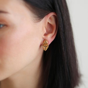 Könnte beinhalten: Goldfarbene Ohrstecker aus Perlen. Die Ohrringe haben eine geometrische, rautenförmige Form und bestehen aus kleinen, gleichmäßig verteilten Perlen. Die Ohrringe werden an einem Ohr vor einem unscharfen Hintergrund gezeigt.