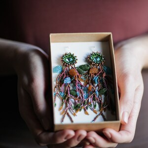 May include: A pair of long, beaded earrings in a box. The earrings are made of brown, green, blue, and orange beads and have a teardrop-shaped design.