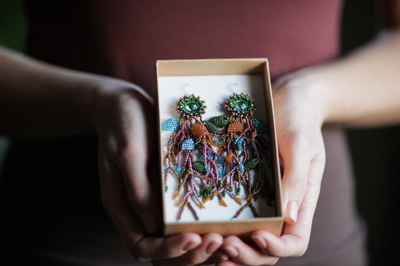 May include: A pair of long, beaded earrings in a box. The earrings are made of brown, green, blue, and orange beads and have a teardrop-shaped design.