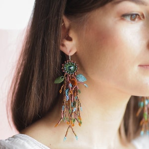 May include: A pair of long, dangling earrings with a floral design. The earrings are made of beads in shades of brown, green, blue, and orange. The earrings are hanging from a woman's ear.