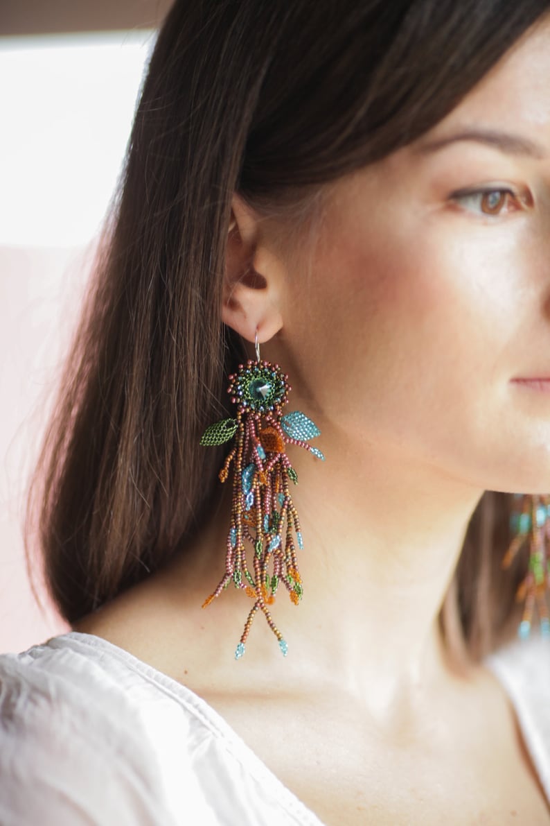 May include: A pair of long, dangling earrings with a floral design. The earrings are made of beads in shades of brown, green, blue, and orange. The earrings are hanging from a woman's ear.