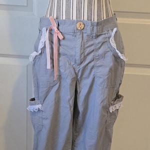 May include: Gray cargo pants with white lace trim on the pockets and a white eyelet fabric skirt. A pink ribbon is tied at the waist. The pants are displayed on a mannequin.