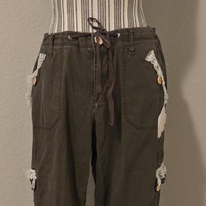 May include: Dark olive green cargo pants with white lace accents and wooden buttons. The pants have a drawstring waist and pockets. The lace is attached to the pockets and sides of the pants.