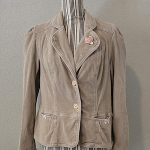 May include: A tan corduroy blazer with a notched collar, two front pockets, and a single-button closure. The blazer features a small pink floral pin and lace cuffs with pink ribbon accents. The jacket is displayed on a mannequin.