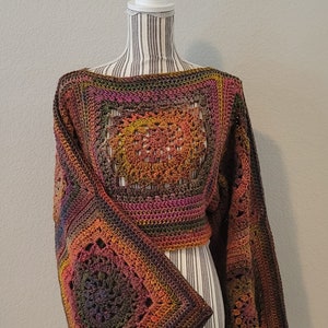 Puede incluir: Un top de crochet con un diseño cuadrado en tonos marrones, naranjas, rosas y verdes. El top tiene mangas largas y anchas y una longitud recortada.