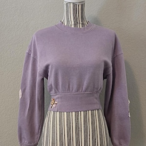 May include: A lilac cropped sweatshirt with long sleeves, shown on a mannequin. The sweatshirt features a ribbed waistband and cuffs. Decorative embellishments are on the sleeves and waistband. The mannequin's torso is covered in a black and white striped fabric.