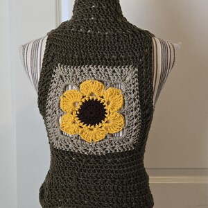 May include: Olive green crochet vest with a high collar. The back features a square design with a yellow sunflower and a dark brown centre, framed by a grey border. The vest is displayed on a striped mannequin.