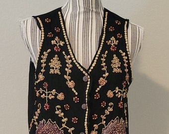 Op de afbeelding: Zwarte vest met bloemenborduurwerk in goud, bordeauxrood en crème. Het mouwloze vest heeft een V-hals, een knoopsluiting en een decoratieve kralenbies. Het vest wordt getoond op een gestreepte paspop.