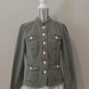 Könnte beinhalten: Olivgrüne Jeansjacke mit Stehkragen, vier Vordertaschen und blütenförmigen Knöpfen. Die Jacke hat weiße Spitzentrimm an den Manschetten und am Saum. Die Jacke wird auf einer Schaufensterpuppe präsentiert.