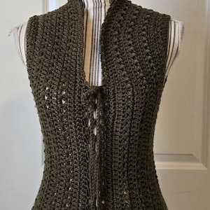 Peut inclure: Gilet en crochet vert olive foncé avec un col montant et une fermeture à nouer. Le gilet fait main est présenté sur un mannequin avec un tissu rayé. Le motif ajouré du gilet est visible.