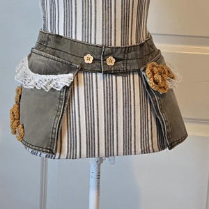 May include: A denim apron with a distressed, olive-green wash. The apron features two pockets with white lace trim and is adorned with crocheted embellishments and flower-shaped buttons. The apron is displayed on a striped mannequin.