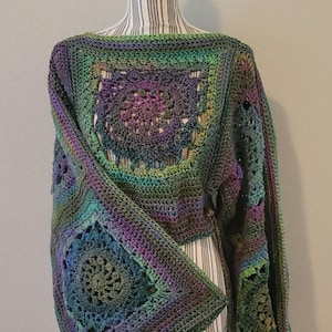 Peut inclure: Un haut crocheté avec un motif carré dans des tons de vert, violet et bleu. Le haut a un col bateau large et des manches longues. Les manches sont évasées et présentent un motif de dentelle crocheté.