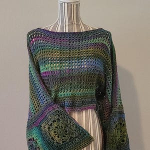 Pode incluir: Um top de crochê multicolorido com mangas sino. O top apresenta uma mistura de azuis, verdes e roxos. As mangas têm um padrão quadrado. O top é exibido em um manequim.