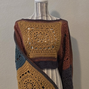 May include: A cropped long-sleeve sweater constructed from multiple crocheted granny squares in shades of mustard yellow, dark brown, charcoal gray, and muted blue. The garment features an open neckline and a boxy silhouette with intricate patterns.