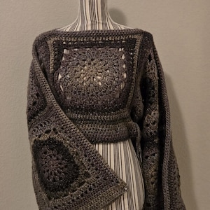 May include: A handmade, dark gray crochet top with a square neckline and long sleeves. The top features a geometric pattern and is displayed on a mannequin with a striped pattern.