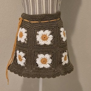 May include: A handmade crochet skirt with a daisy pattern. The skirt has a dark olive green base with white and yellow daisy appliqués. It features a scalloped hem and a mustard yellow drawstring.