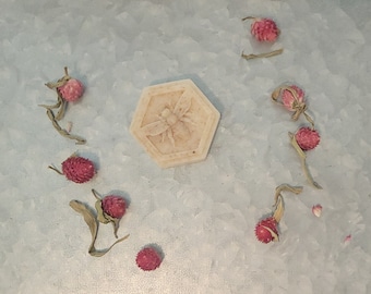 May include: A hexagonal shaped soap bar with a bee design in the center. The soap is surrounded by dried pink flowers on a gray background.