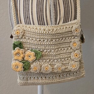 May include: A beige crochet crossbody bag with a floral design. The bag features a rectangular shape with a flap closure and a long strap. The flowers are yellow and white with green leaves, adding a decorative touch to the handmade bag.