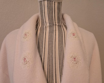 Könnte beinhalten: Ein blassrosa Mantel mit einem breiten Schalkragen, der mit vier kreisförmigen cremefarbenen Spitzenapplikationen verziert ist. Jedes Spitzendetail enthält einen kleinen, mittigen rosa Blumenakzent. Der Mantel wird auf einer schwarz-weiß gestreiften Schneiderpuppe präsentiert.