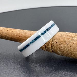 May include: White ceramic ring with a thin blue line inlay. The ring is on a wooden surface.