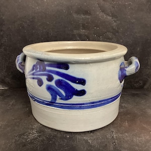 May include: A white ceramic crock with blue and white floral designs and two handles. The crock is a traditional German style.