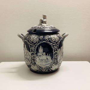 May include: A blue and white ceramic pitcher with a lid and a handle on each side. The pitcher is decorated with a white castle and grapevines.