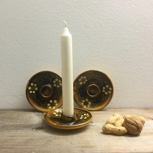 May include: A white candle stands in a brown ceramic candle holder with floral accents. Two additional matching candle holders are behind it. The candle holders have a dark brown center and a lighter brown rim with white and yellow floral designs.